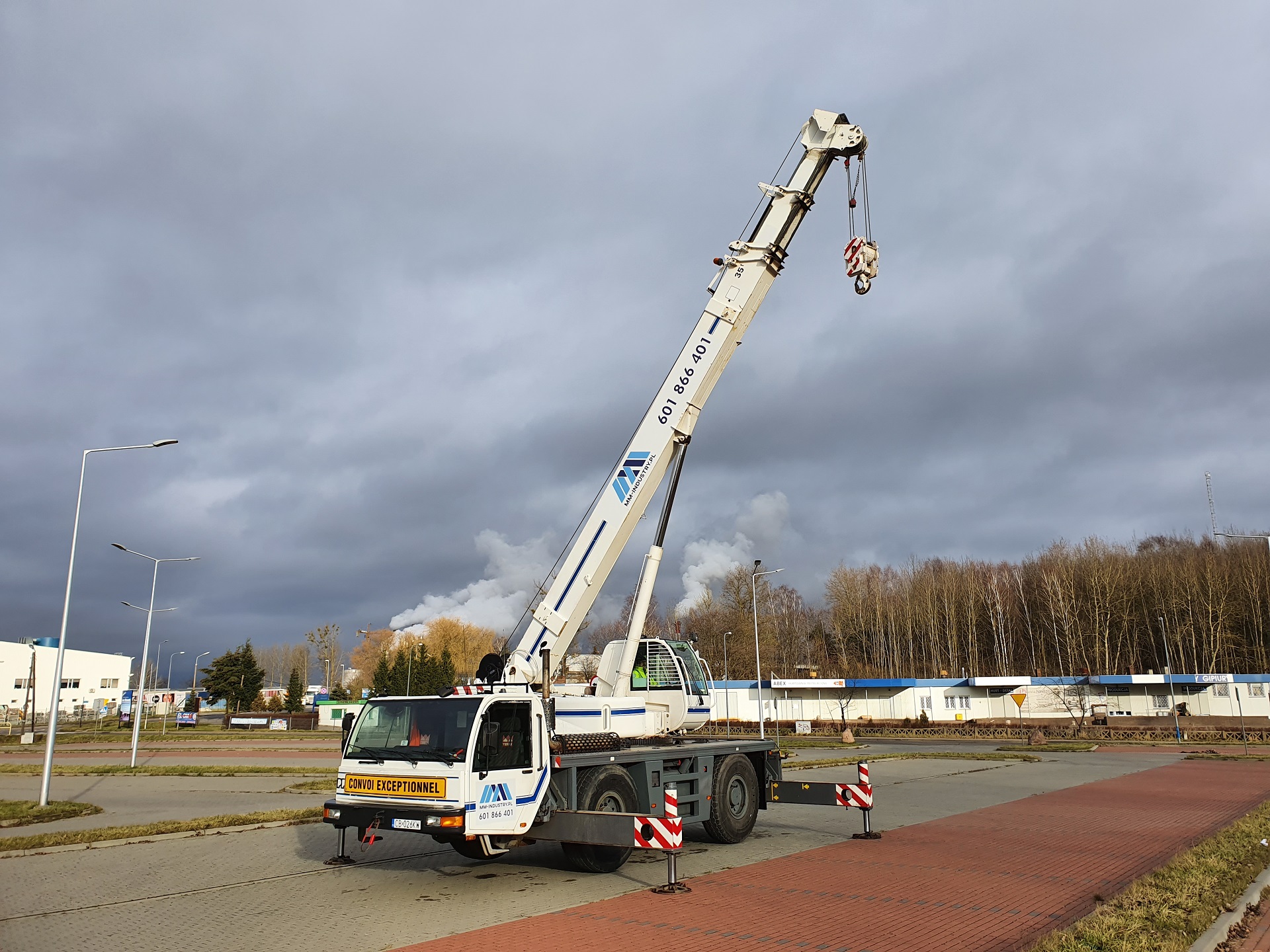 Żuraw samochodowy z wysuniętym ramieniem na tle pochmurnego nieba i zabudowań przemysłowych, z widocznym napisem 'Convoi Exceptionnel' na kabinie.