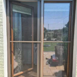 Zewnętrzne drzwi przesuwne z moskitierą w brązowej ramie, jedno skrzydło otwarte, widok na ogród.