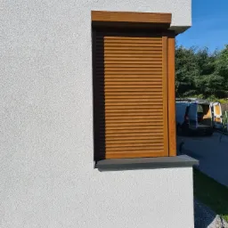 Zamknięta, drewniana roleta zewnętrzna na tle białej, teksturowanej ściany domu, z widocznym samochodem dostawczym w tle.