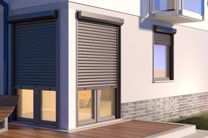 Elewacja domu z oknami i drzwiami balkonowymi zabezpieczonymi szarymi roletami zewnętrznymi, widok z drewnianego tarasu.