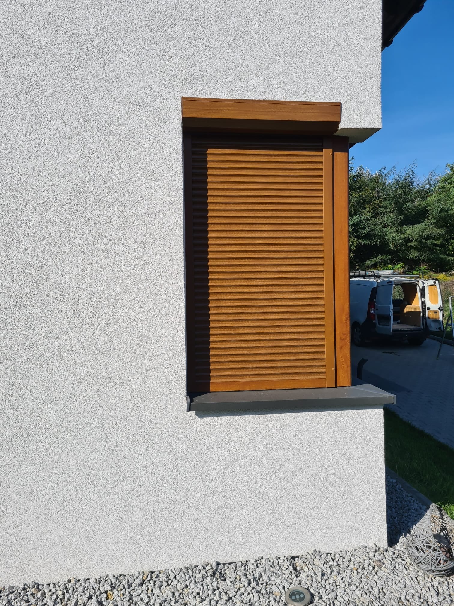 Zamknięta, drewniana roleta zewnętrzna na tle białej, teksturowanej ściany domu, z widocznym samochodem dostawczym w tle.