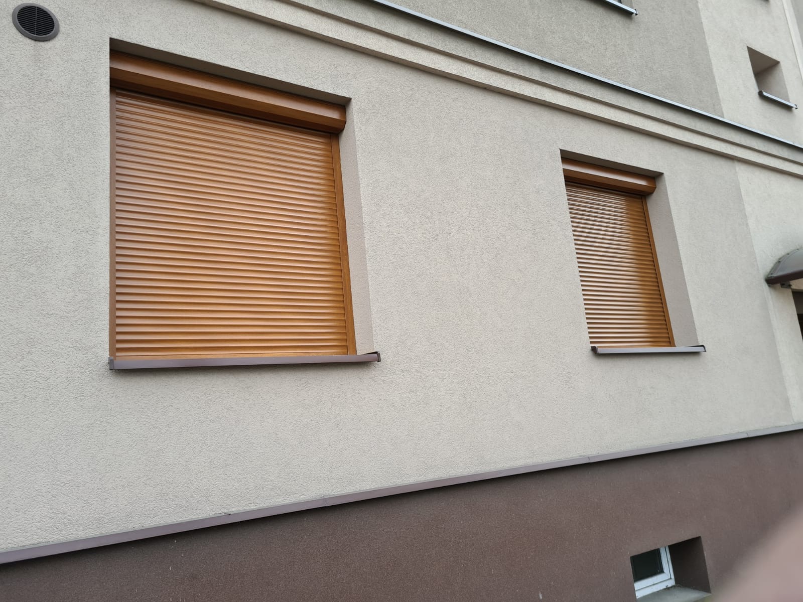 Dwa okna z brązowymi roletami zewnętrznymi na elewacji budynku z jasnoszarym tynkiem strukturalnym i ciemnobrązowym cokołem.