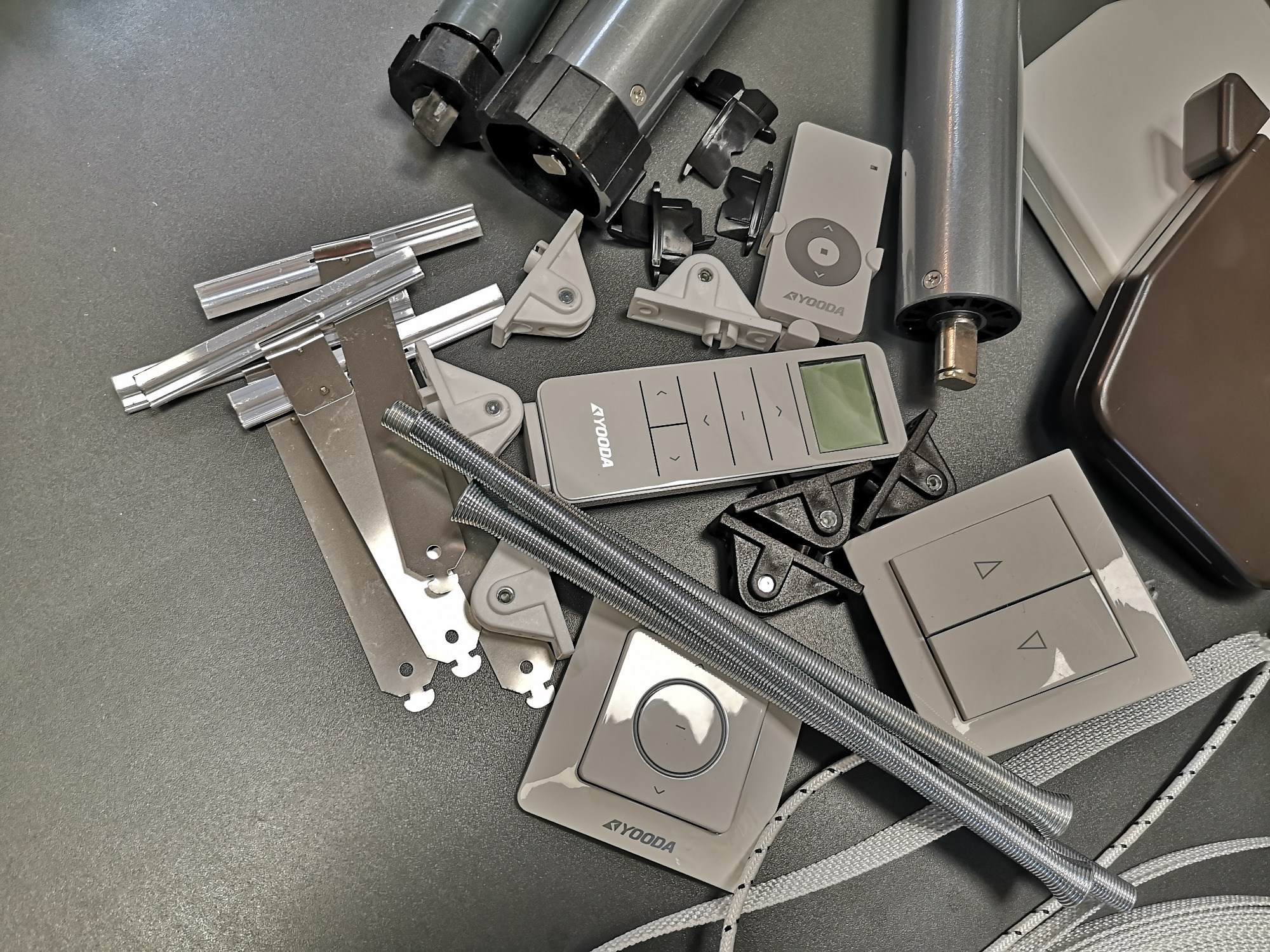 Elementy montażowe do rolet: silniki, piloty, przełączniki, sprężyny i uchwyty na szarym tle, marki Ayooda.