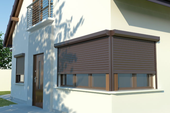 Brązowa roleta zewnętrzna całkowicie zasłonięta na oknie domu z białymi ścianami, widoczne cienie drzew na elewacji.