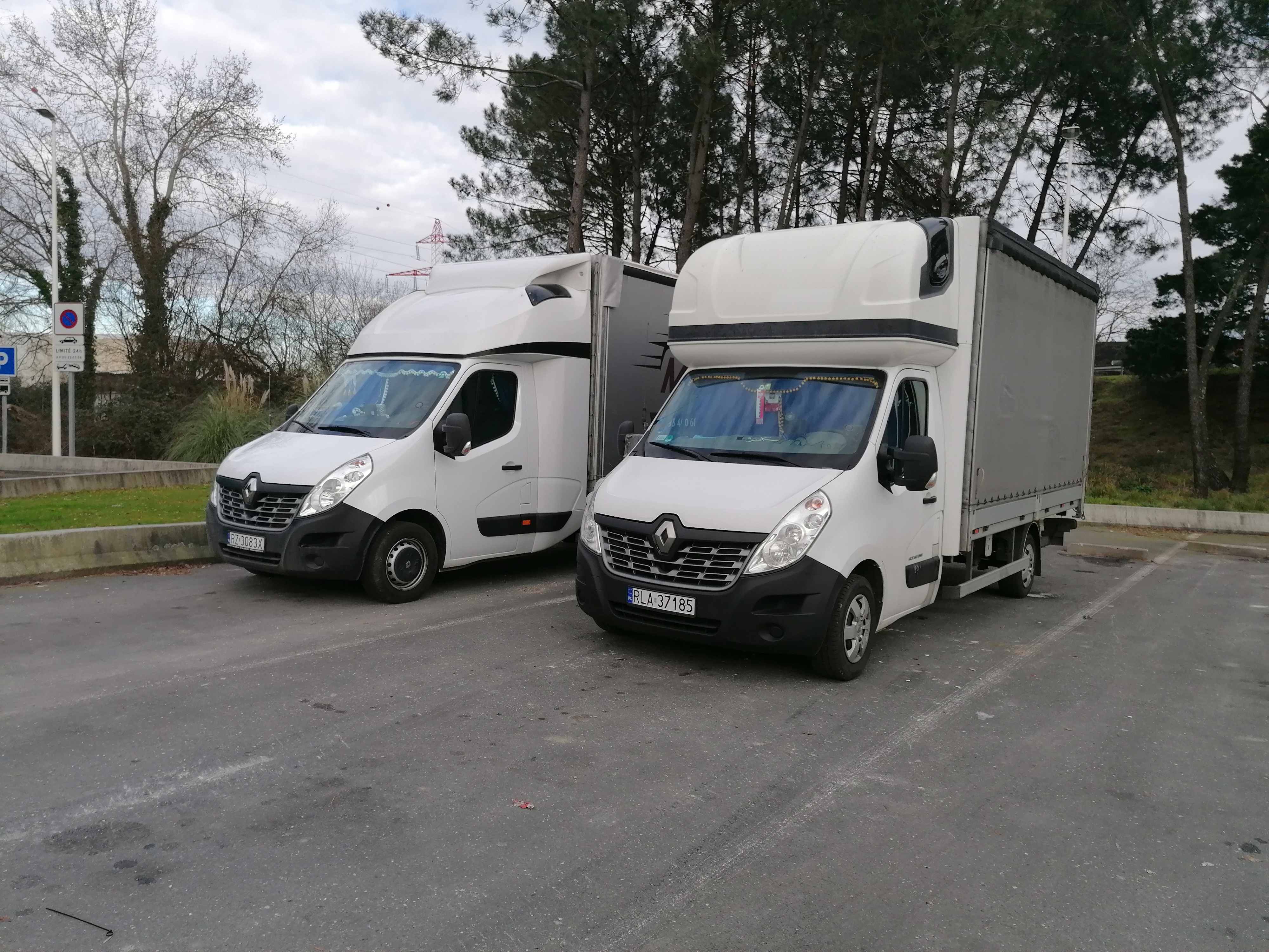 Dwa białe busy Renault Master z plandekami na parkingu, jeden z dekoracjami na przedniej szybie, drzewa w tle.