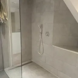 Nowoczesna kabina prysznicowa z szarymi, prostokątnymi płytkami, deszczownicą i panelem sterującym, widoczny odpływ liniowy i półka w ścianie, w tle drzwi do innego pomieszczenia.