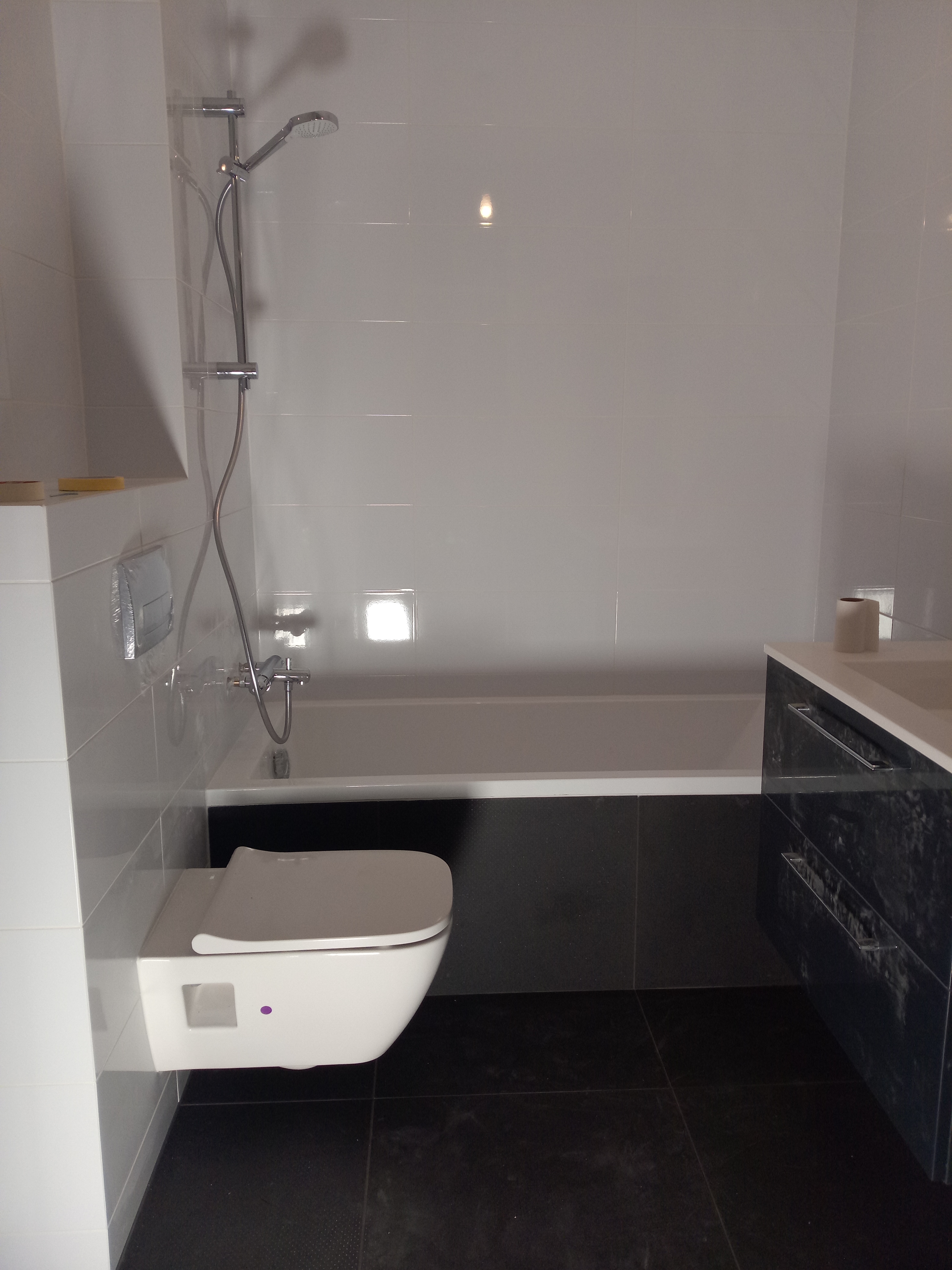 Nowoczesna łazienka z białymi płytkami ściennymi, wiszącą toaletą z fioletowym akcentem, czarną podłogą, wanną i umywalką z szafką o marmurkowym wzorze.