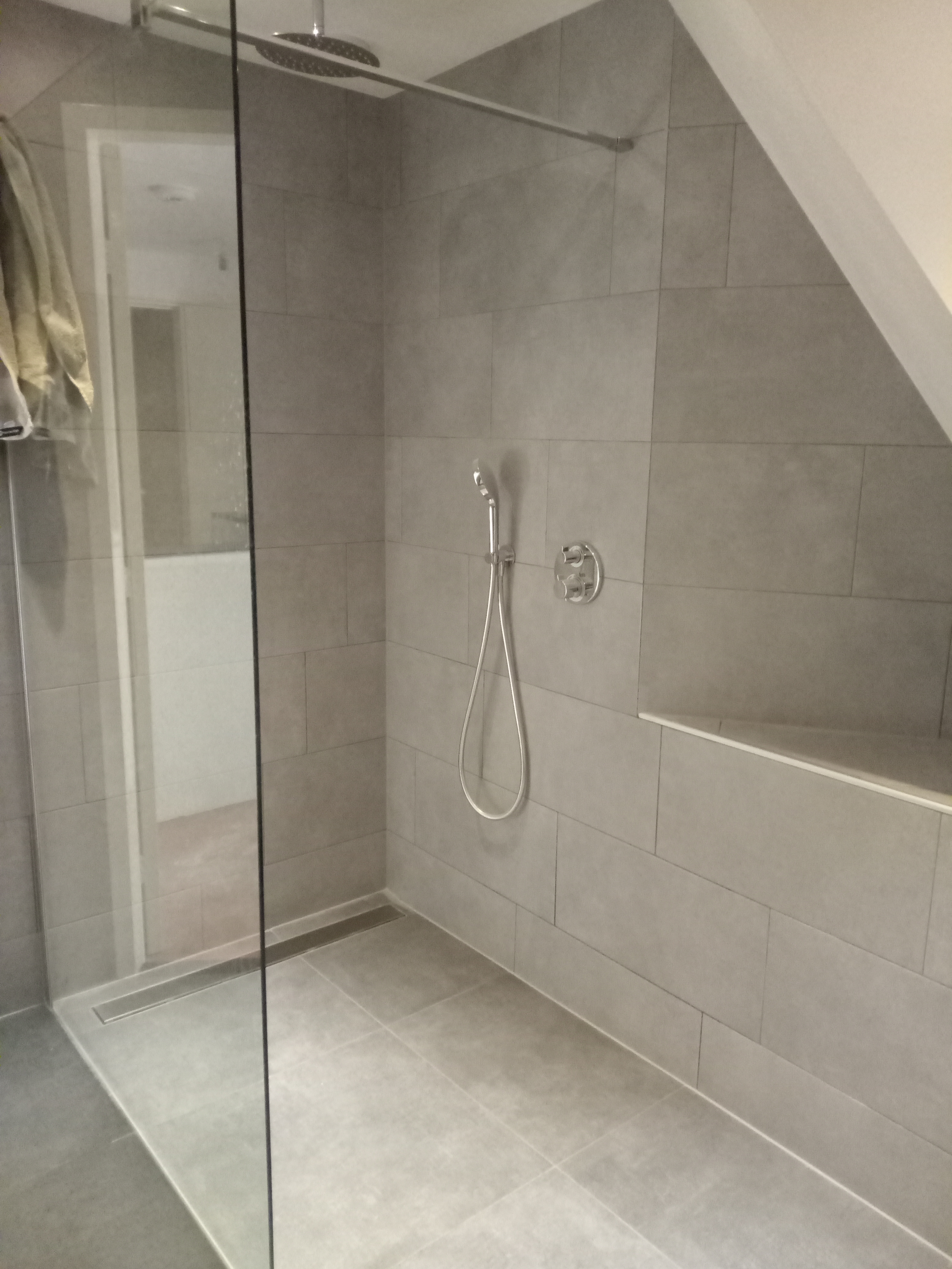 Nowoczesna kabina prysznicowa z szarymi, prostokątnymi płytkami, deszczownicą i panelem sterującym, widoczny odpływ liniowy i półka w ścianie, w tle drzwi do innego pomieszczenia.