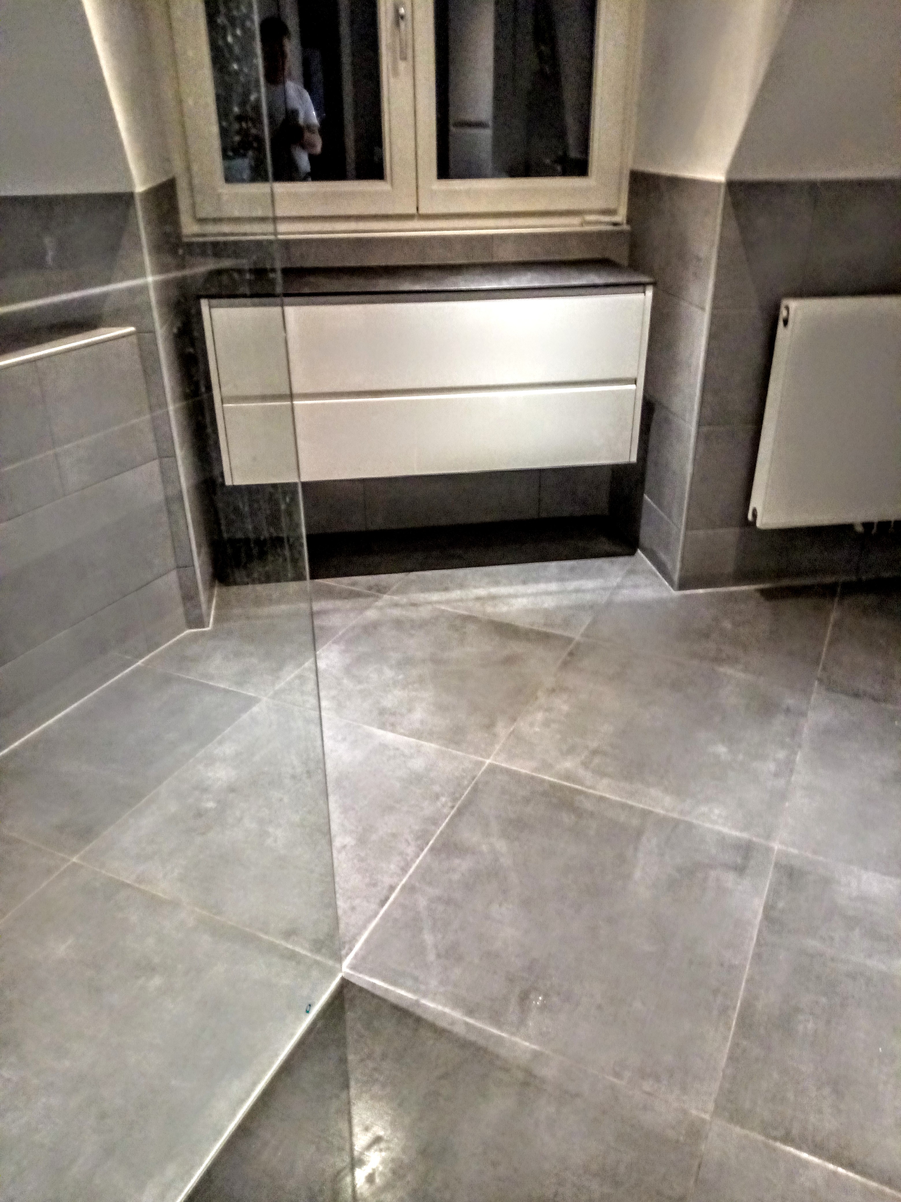 Wykończona łazienka z szarymi płytkami podłogowymi i ściennymi, białą szafką pod umywalką i grzejnikiem na ścianie, widoczny fragment szklanej kabiny prysznicowej.