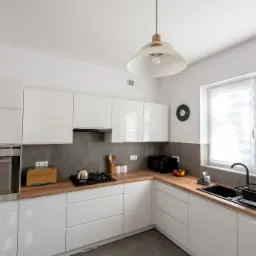 Jasna kuchnia z białymi, lakierowanymi frontami, drewnianym blatem, szarym pasem nad blatem, czarnym zlewem i osprzętem oraz lampą wiszącą w stylu retro.