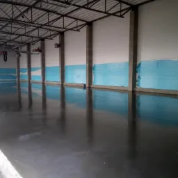 Wnętrze hali produkcyjnej z nową, błyszczącą, ciemnoszarą posadzką przemysłową; ściany częściowo zabezpieczone folią ochronną.