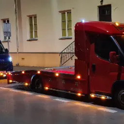 Dwa samochody lawety Iveco w kolorze czerwonym i niebieskim, zaparkowane nocą na ulicy przed budynkiem, z włączonymi światłami obrysowymi i reflektorami.