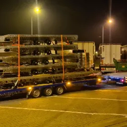 Ciężarówka Iveco z naczepą przewożąca kilka metalowych konstrukcji na parkingowej przestrzeni oświetlonej latarniami ulicznymi, w tle inne pojazdy ciężarowe.