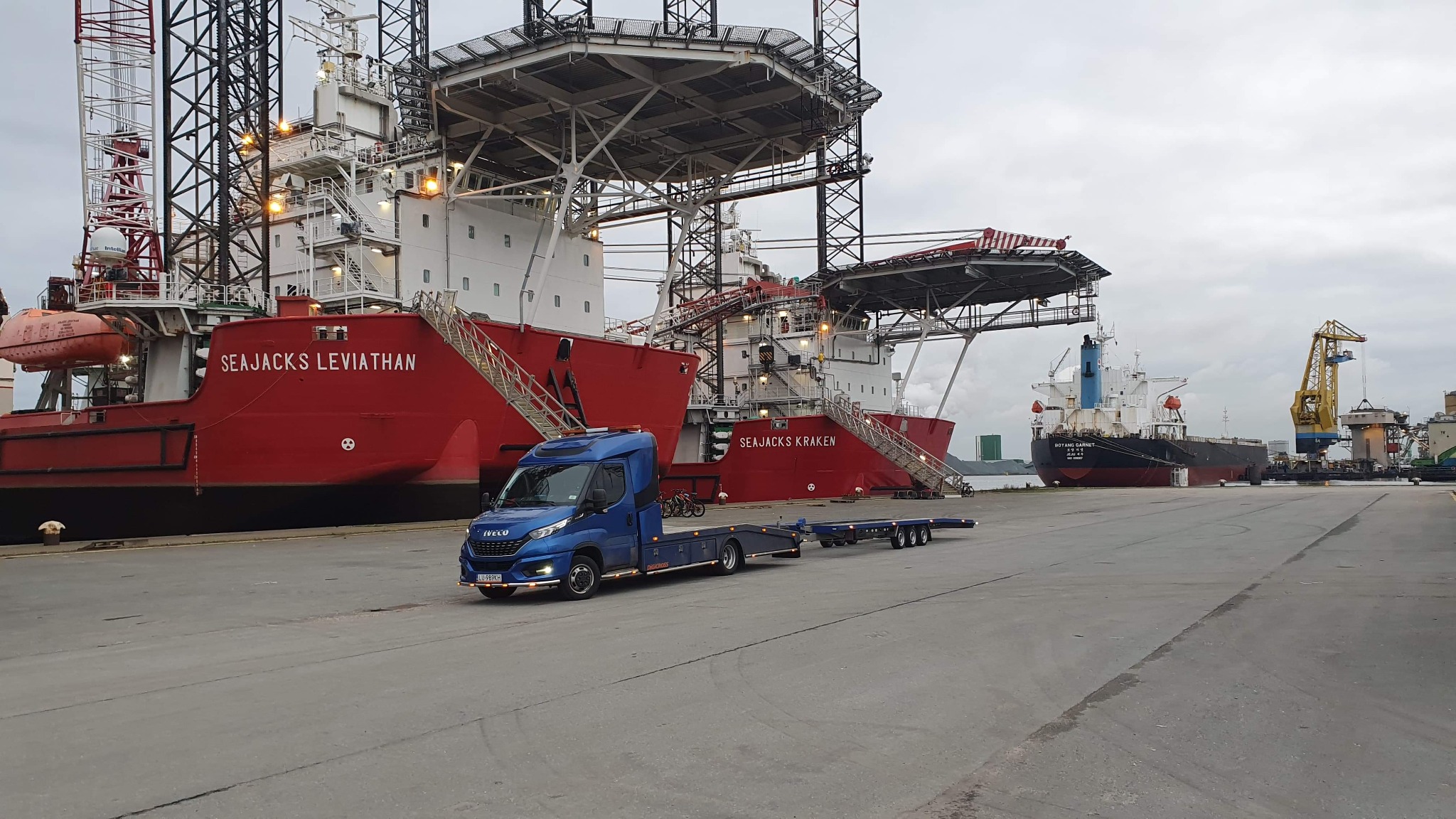 Niebieska laweta Iveco z przyczepą na tle dwóch czerwonych statków Sea Jacks Leviathan i Sea Jacks Kraken w porcie.