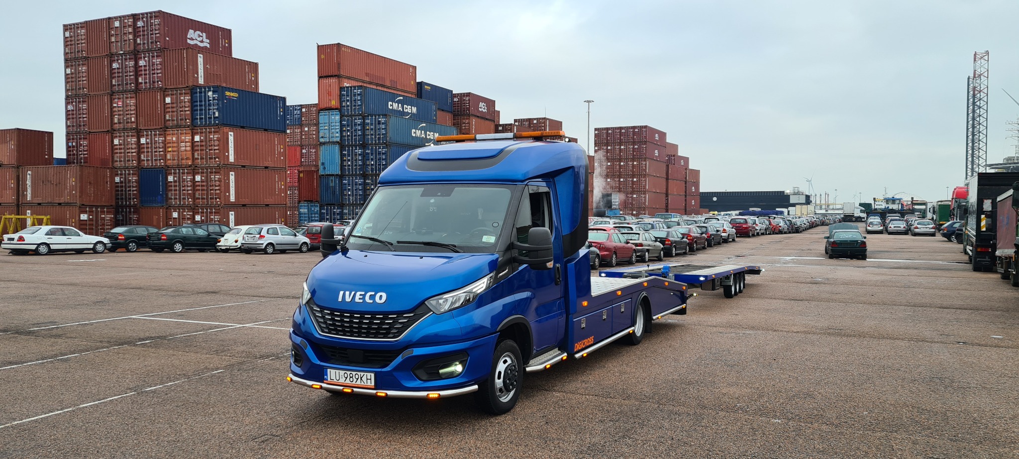Niebieska laweta IVECO na placu z rzędami samochodów i kontenerami w tle, widoczne detale takie jak tablica rejestracyjna i dodatkowe oświetlenie
