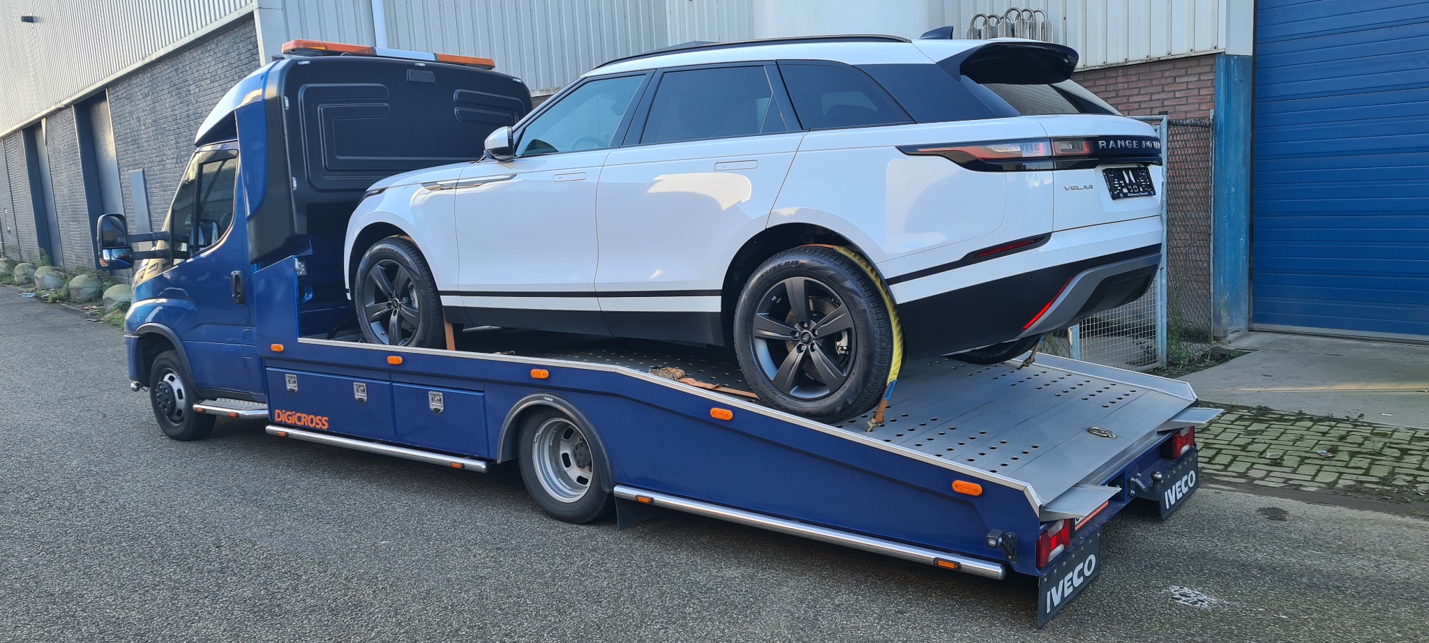Biały Range Rover Velar zabezpieczony pasami na platformie lawety Digicross Iveco, widok z tyłu, na tle szarego budynku.
