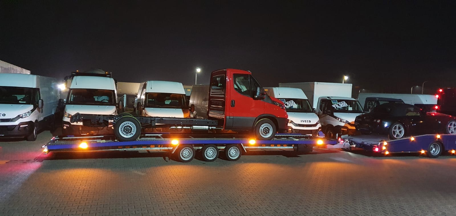Czerwony Iveco Daily bez zabudowy na niskopodwoziowej lawecie, obok białe furgonetki Iveco i czarny kabriolet Mazda MX-5, nocne oświetlenie placu.
