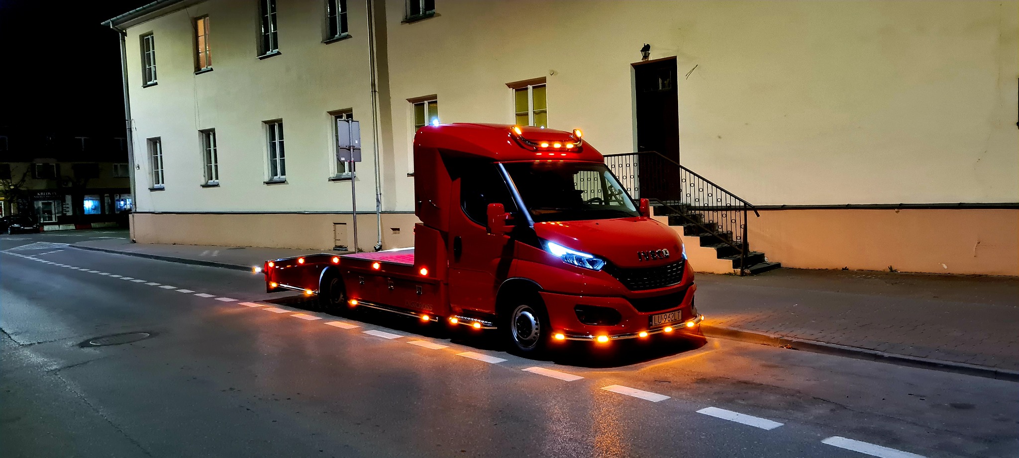 Czerwona laweta Iveco z włączonymi światłami pozycyjnymi stoi na ulicy w Łukowie na tle budynku oświetlonego lampami ulicznymi.