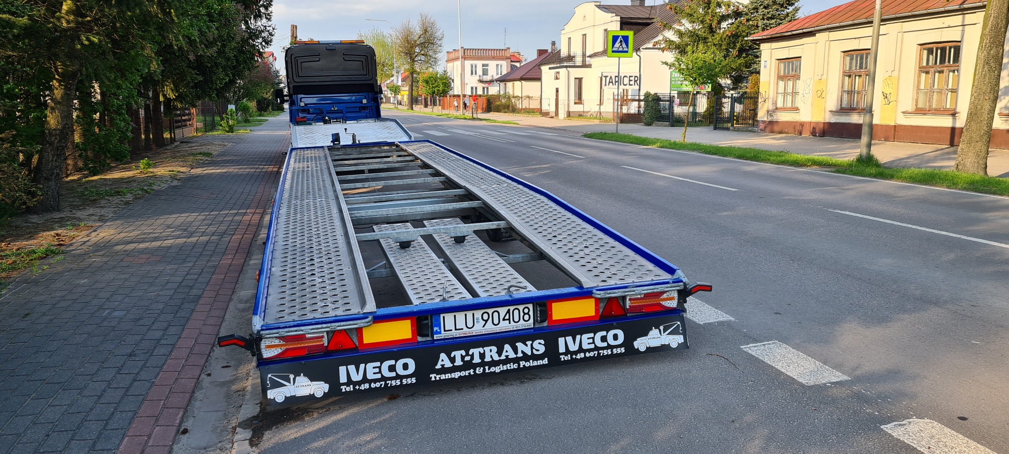 Pusta, niebieska laweta samochodowa marki Iveco, zaparkowana na poboczu ulicy w słoneczny dzień, z widocznymi budynkami w tle.