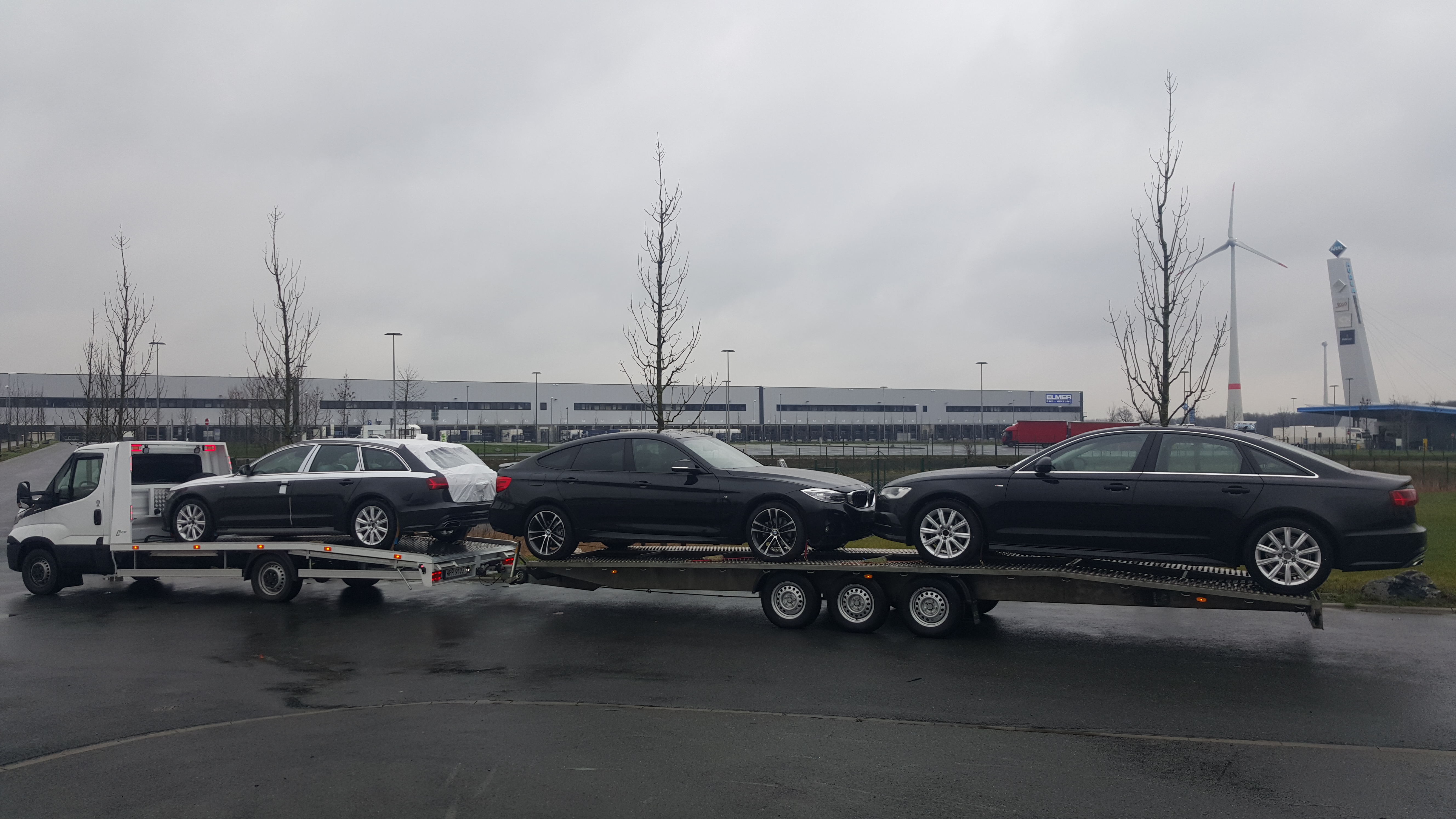 Laweta z trzema czarnymi samochodami osobowymi na tle szarego nieba i budynku magazynowego z wiatrakiem w oddali.