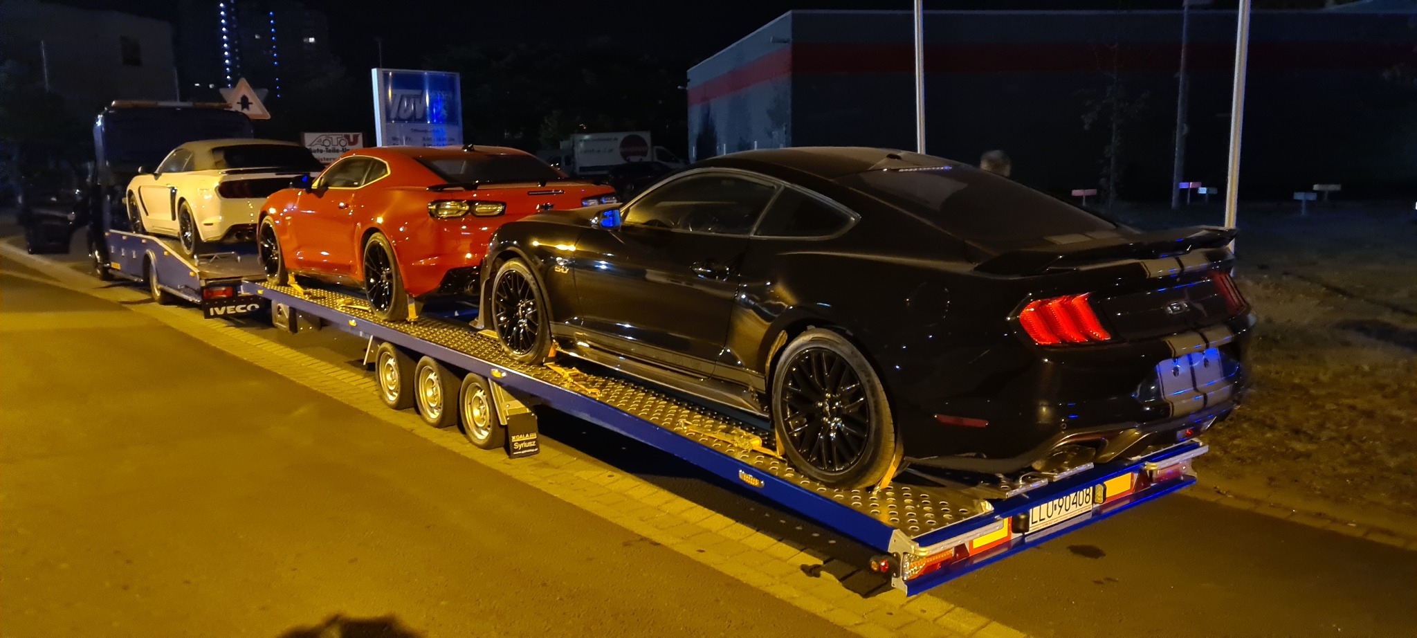 Laweta z trzema sportowymi samochodami: białym kabrioletem, czerwonym coupe i czarnym Mustangiem z szarymi pasami, widziana nocą na parkingu.