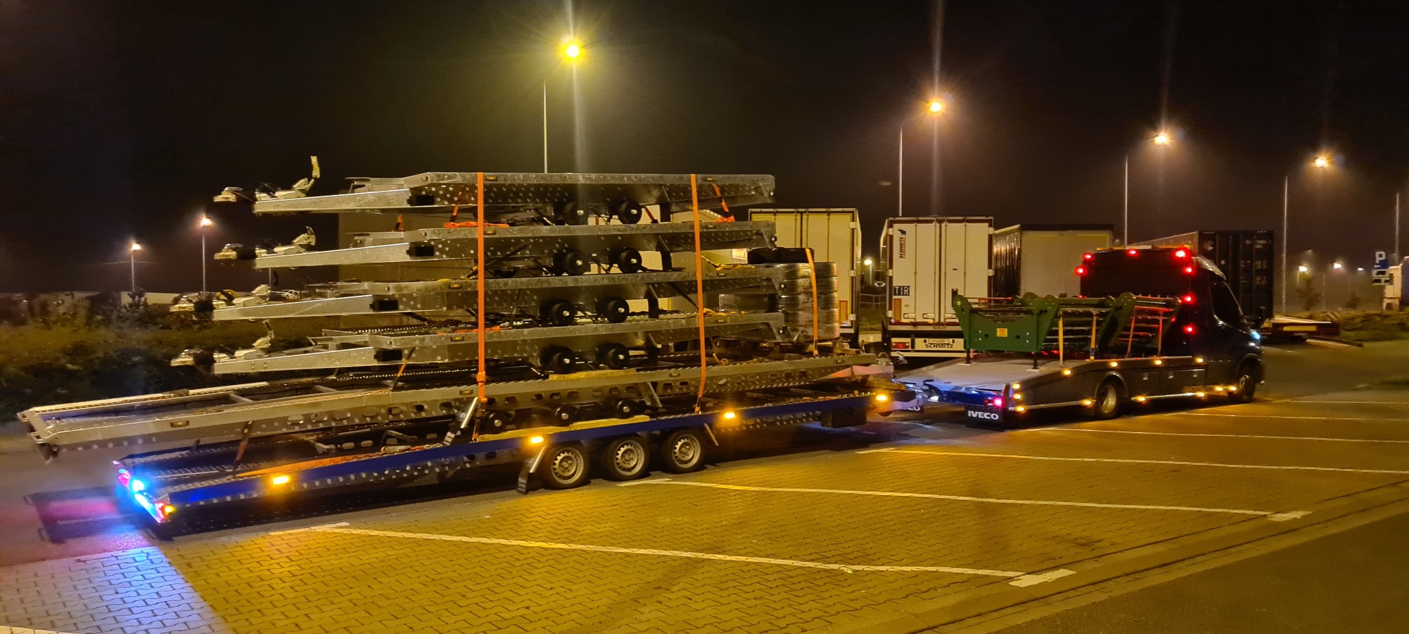 Ciężarówka Iveco z naczepą przewożąca kilka metalowych konstrukcji na parkingowej przestrzeni oświetlonej latarniami ulicznymi, w tle inne pojazdy ciężarowe.