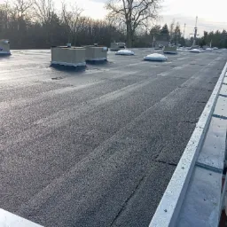 Płaski dach pokryty papą termozgrzewalną z widocznymi świetlikami, kominkami wentylacyjnymi i obróbkami blacharskimi przy krawędzi, zabezpieczony barierkami na rusztowaniu.