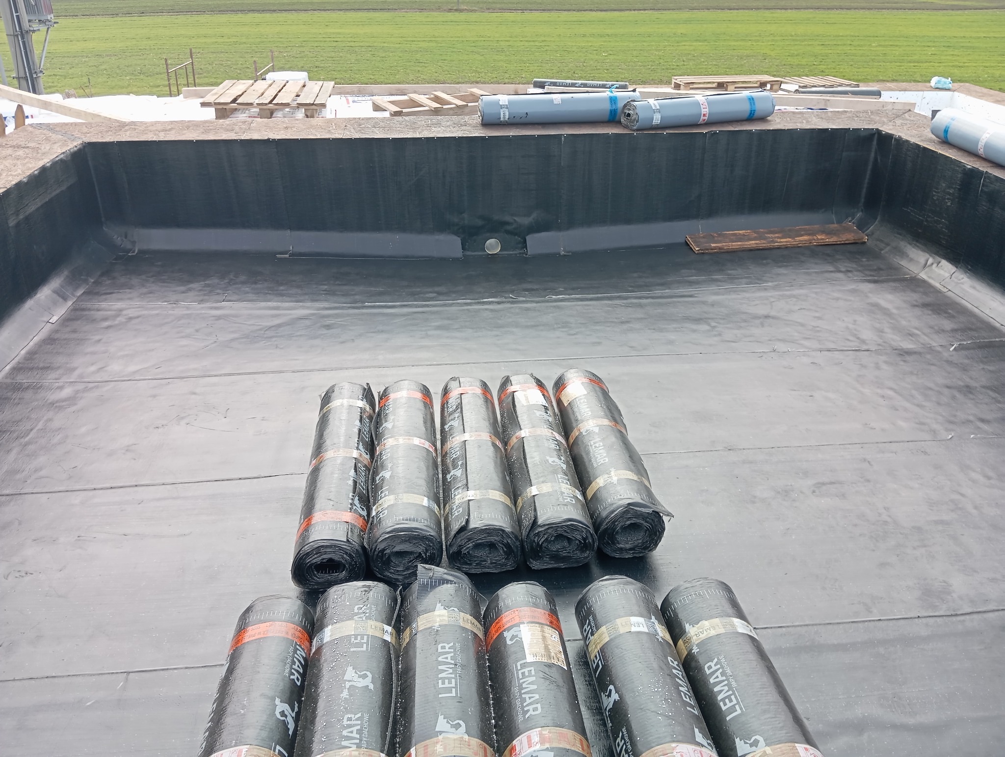 Hydroizolacja dachu płaskiego w trakcie realizacji, widoczne zrolowane arkusze papy dachowej Lemar na powierzchni pokrytej czarną membraną, w tle zielone pole.