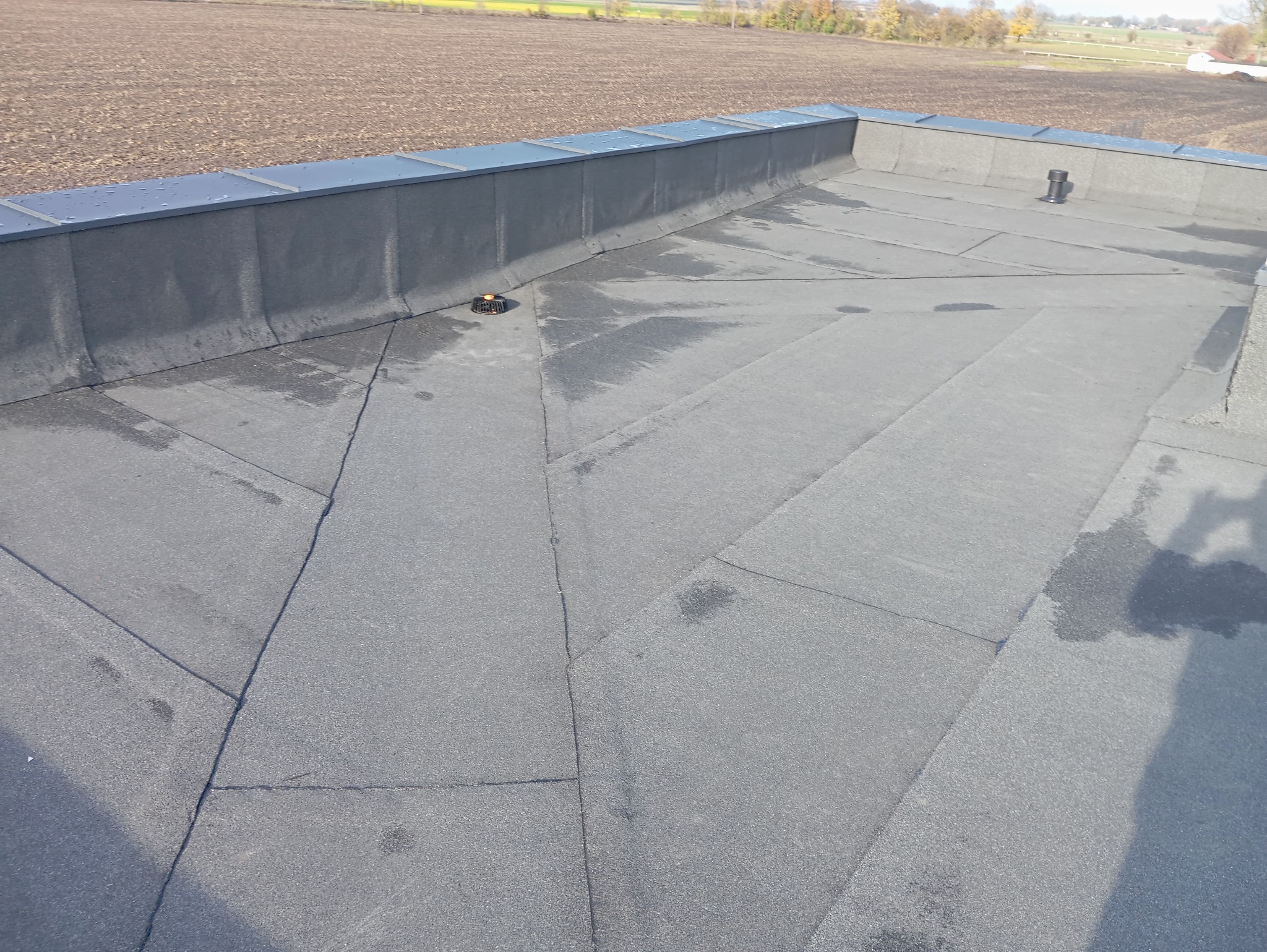 Płaski dach pokryty papą termozgrzewalną, z widocznym kominkiem wentylacyjnym i obróbkami blacharskimi na krawędzi, na tle zaoranego pola.