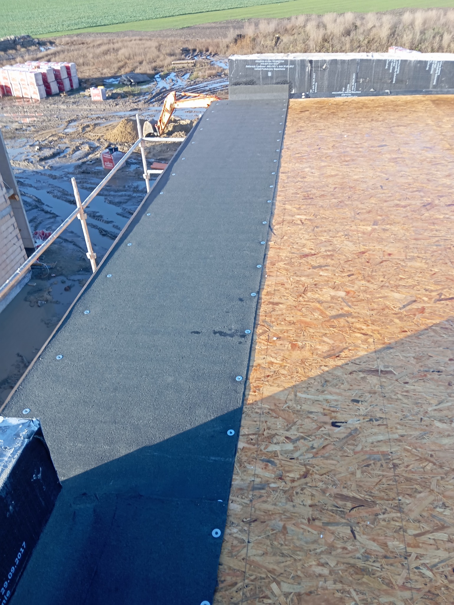 Widok z góry na fragment dachu w trakcie prac: po lewej czarny pas papy mocowany do drewnianej konstrukcji z płyty OSB za pomocą białych podkładek, w tle rusztowanie i materiały budowlane.