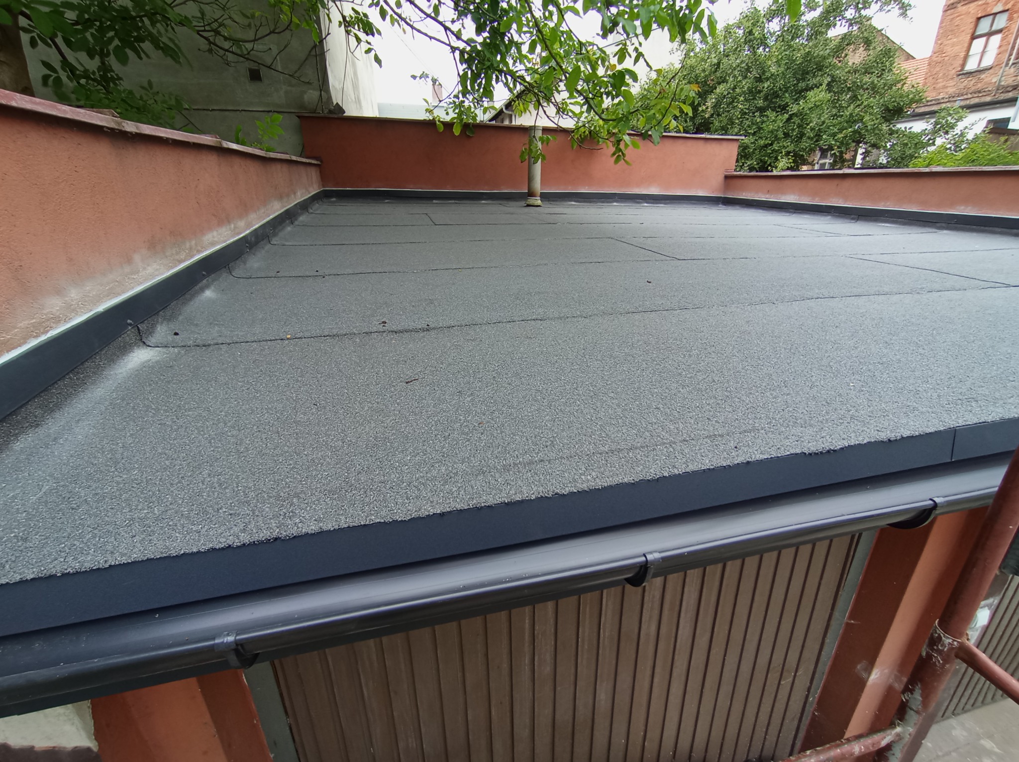 Widok z góry na płaski dach pokryty papą termozgrzewalną, z widocznymi czarnymi rynnami i fragmentem ściany budynku w tle, częściowo przysłonięty gałęziami drzewa.