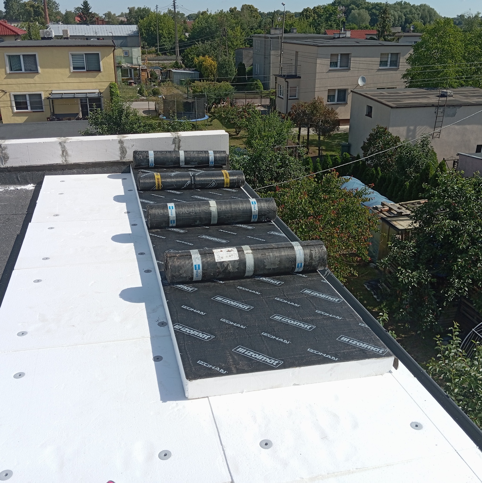 Układanie izolacji termicznej z płyt styropianowych i papy termozgrzewalnej na dachu płaskim, widoczne rolki papy Izohan przygotowane do montażu.