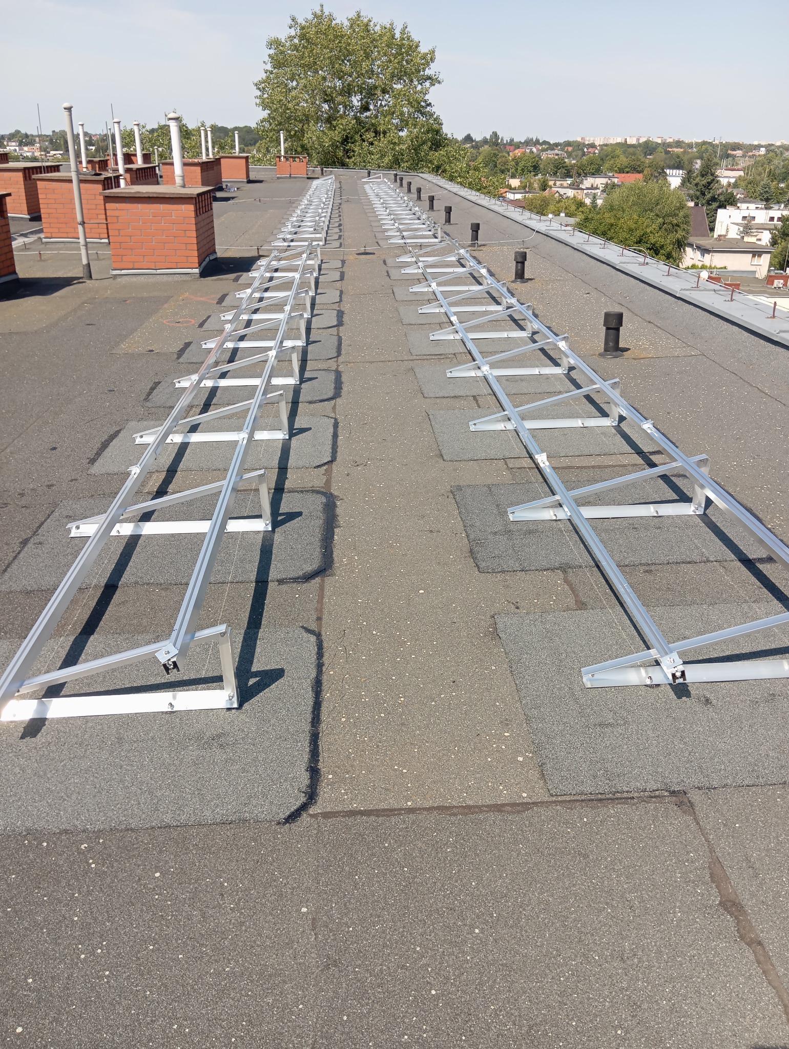 Montaż aluminiowej konstrukcji wsporczej na płaskim dachu pokrytym papą, przygotowanie do instalacji paneli słonecznych, widoczne kominy i zabudowa miejska w tle.