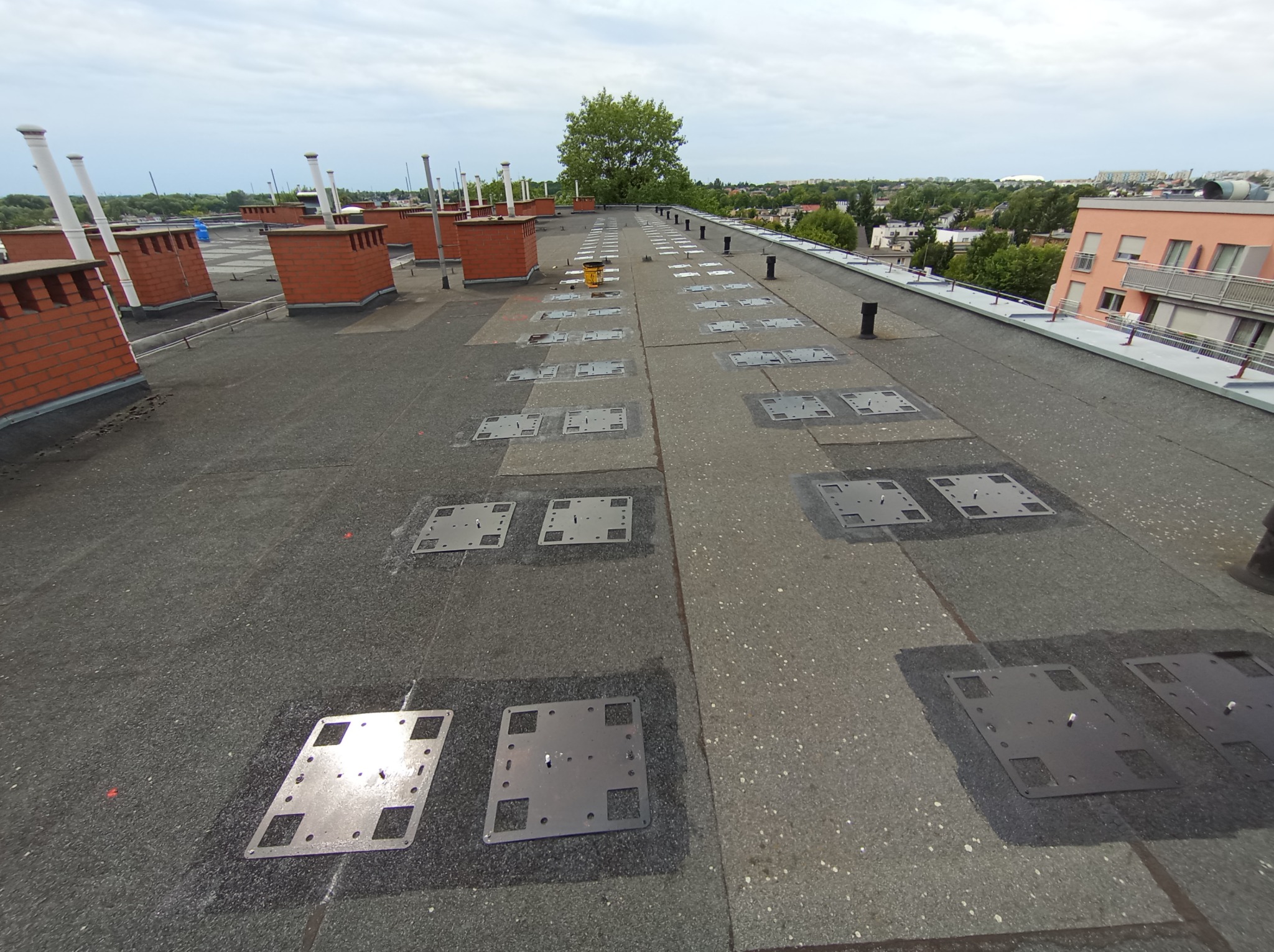 Płaski dach pokryty papą, z zamontowanymi metalowymi podstawami pod instalacje, widoczne kominy wentylacyjne z cegły i otoczenie miejskie w tle.