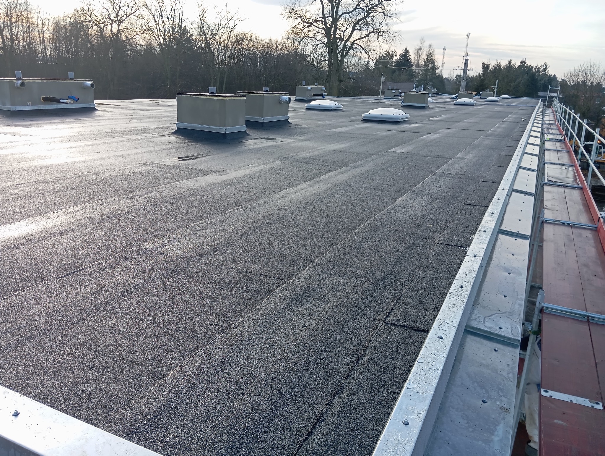 Płaski dach pokryty papą termozgrzewalną z widocznymi świetlikami, kominkami wentylacyjnymi i obróbkami blacharskimi przy krawędzi, zabezpieczony barierkami na rusztowaniu.