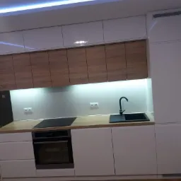 Nowoczesna kuchnia z białymi szafkami dolnymi i górnymi na wysoki połysk oraz drewnopodobnymi szafkami wiszącymi, czarnym zlewem i płytą indukcyjną, oświetlona listwą LED pod szafkami.