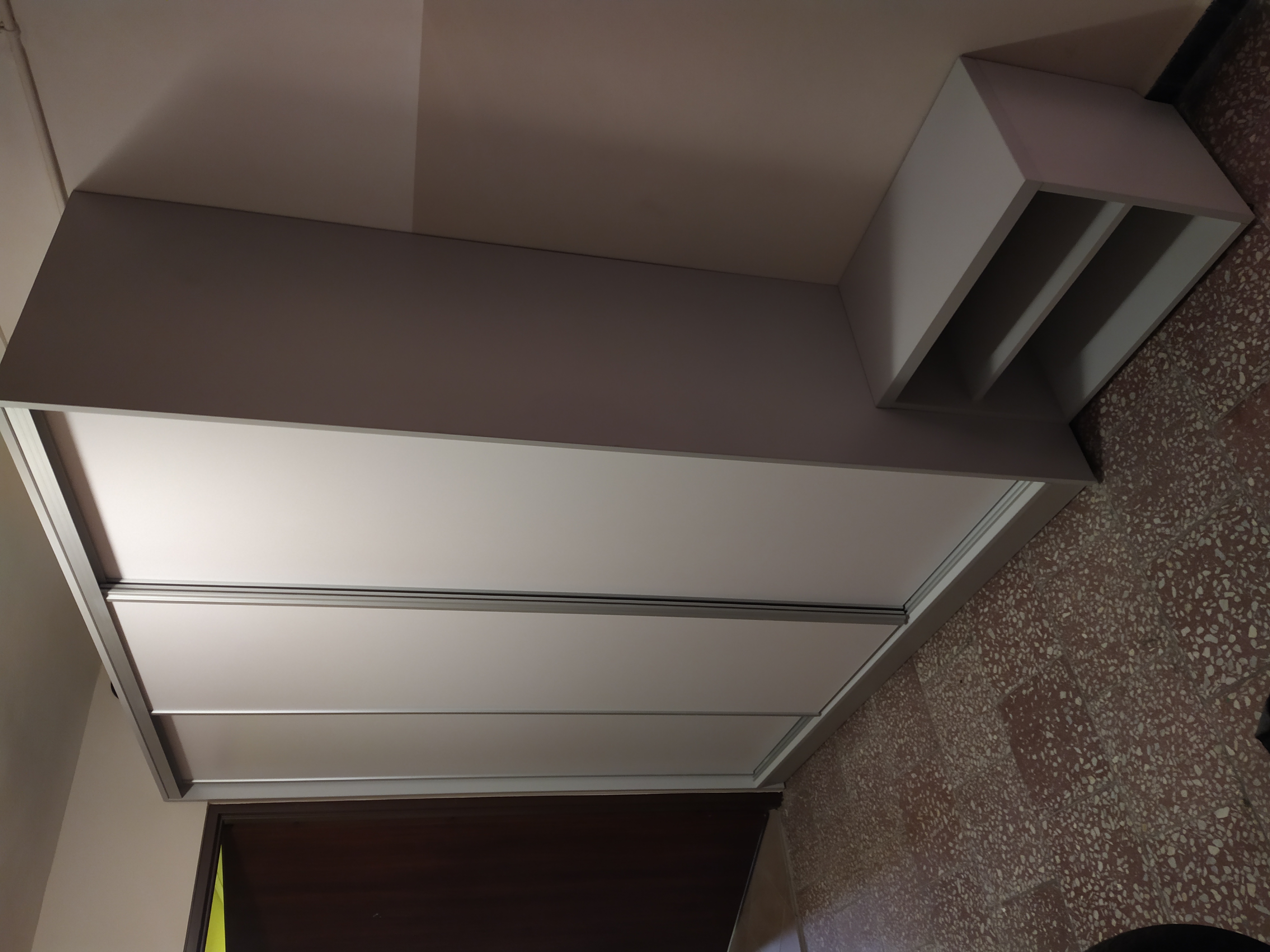 Jasnoszara szafa przesuwna z nadstawką z półkami, sfotografowana z dołu, ukazująca strukturę terazzo na podłodze.