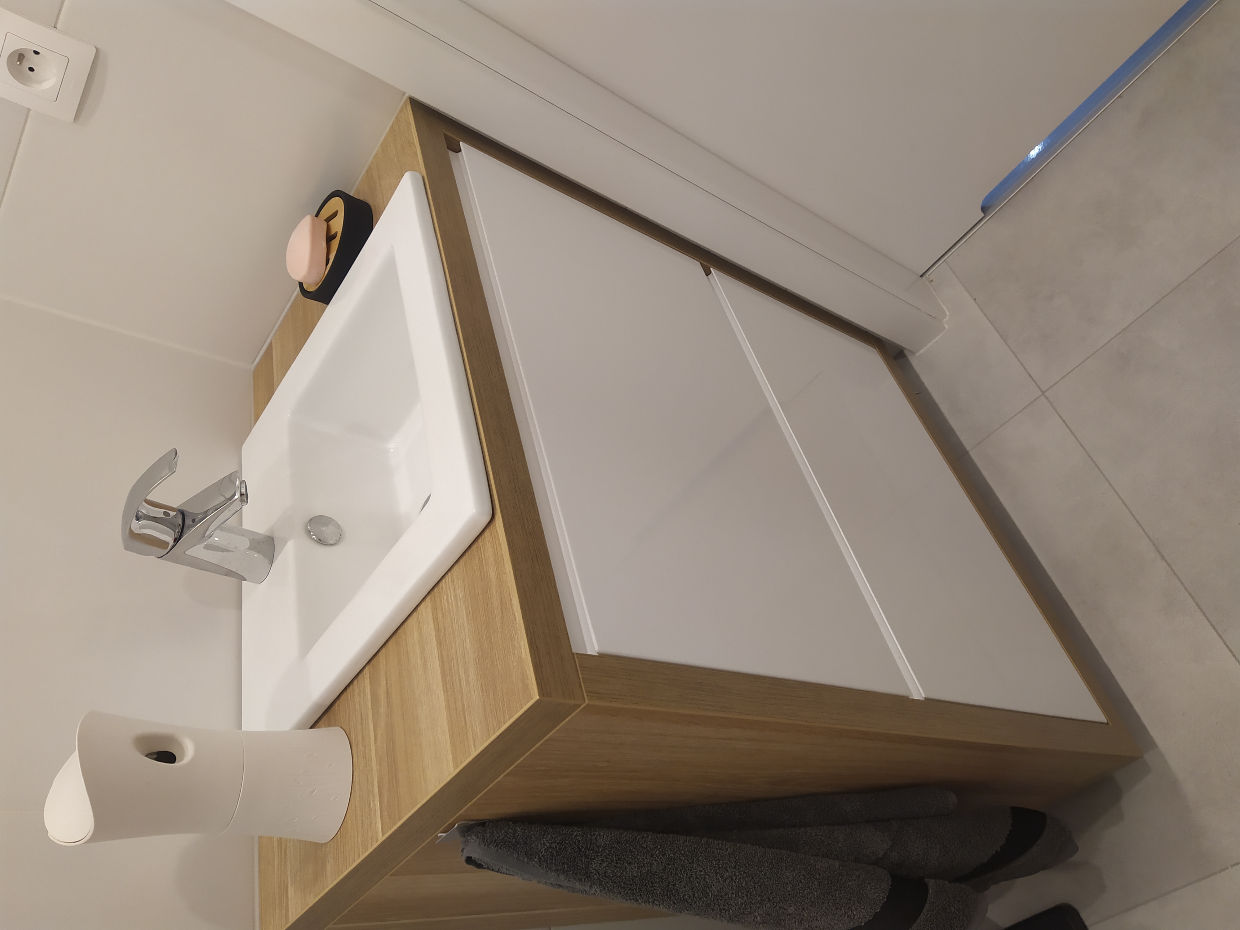 Biała umywalka z chromowaną baterią, osadzona na szafce z drewnianym blatem i białymi frontami, uzupełniona o dozownik mydła i ręczniki w łazience z szarą płytką podłogową.