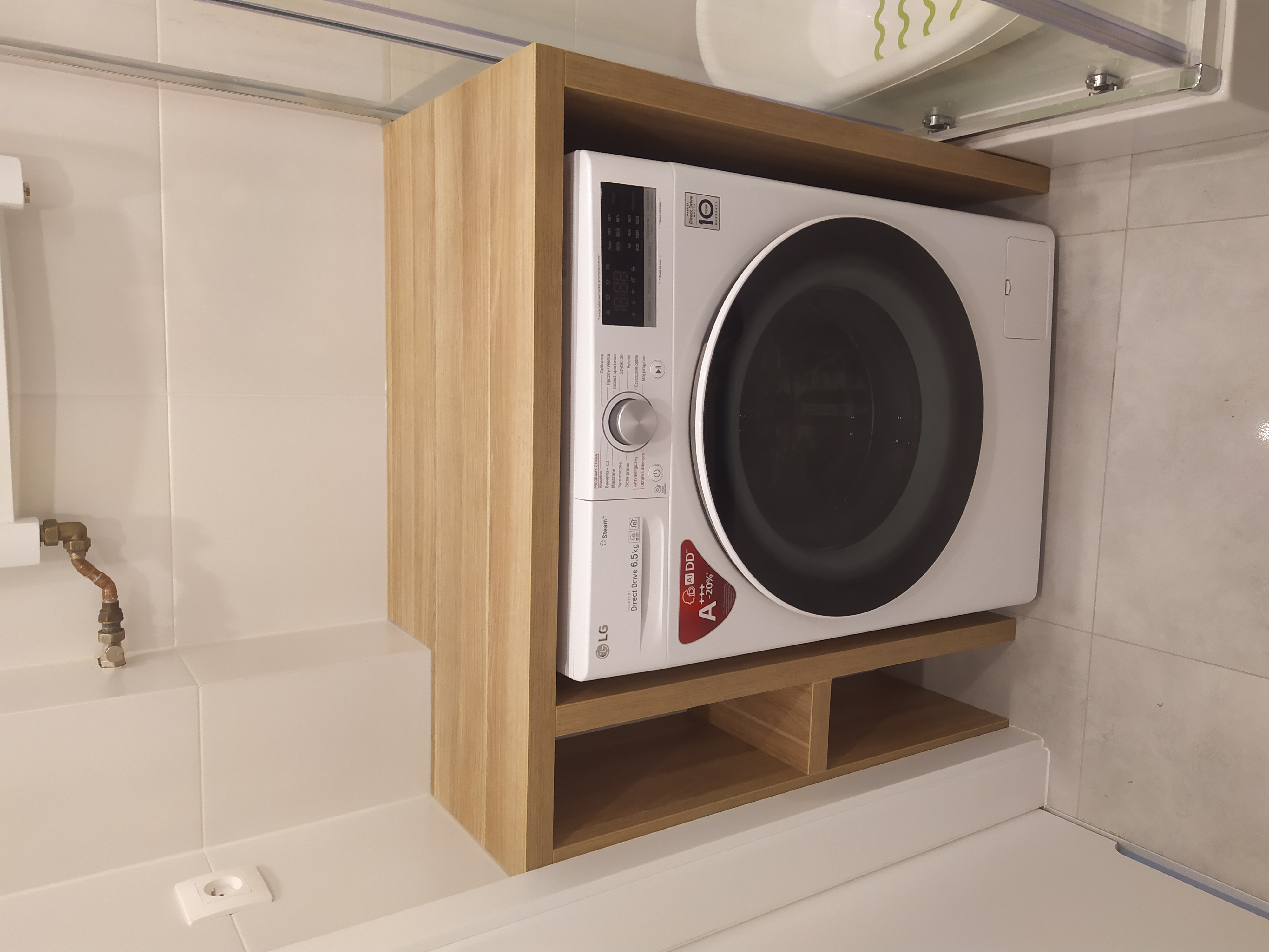 Biała pralka LG Direct Drive 6.5kg umieszczona w drewnianej obudowie z półkami w łazience wyłożonej jasnymi płytkami.
