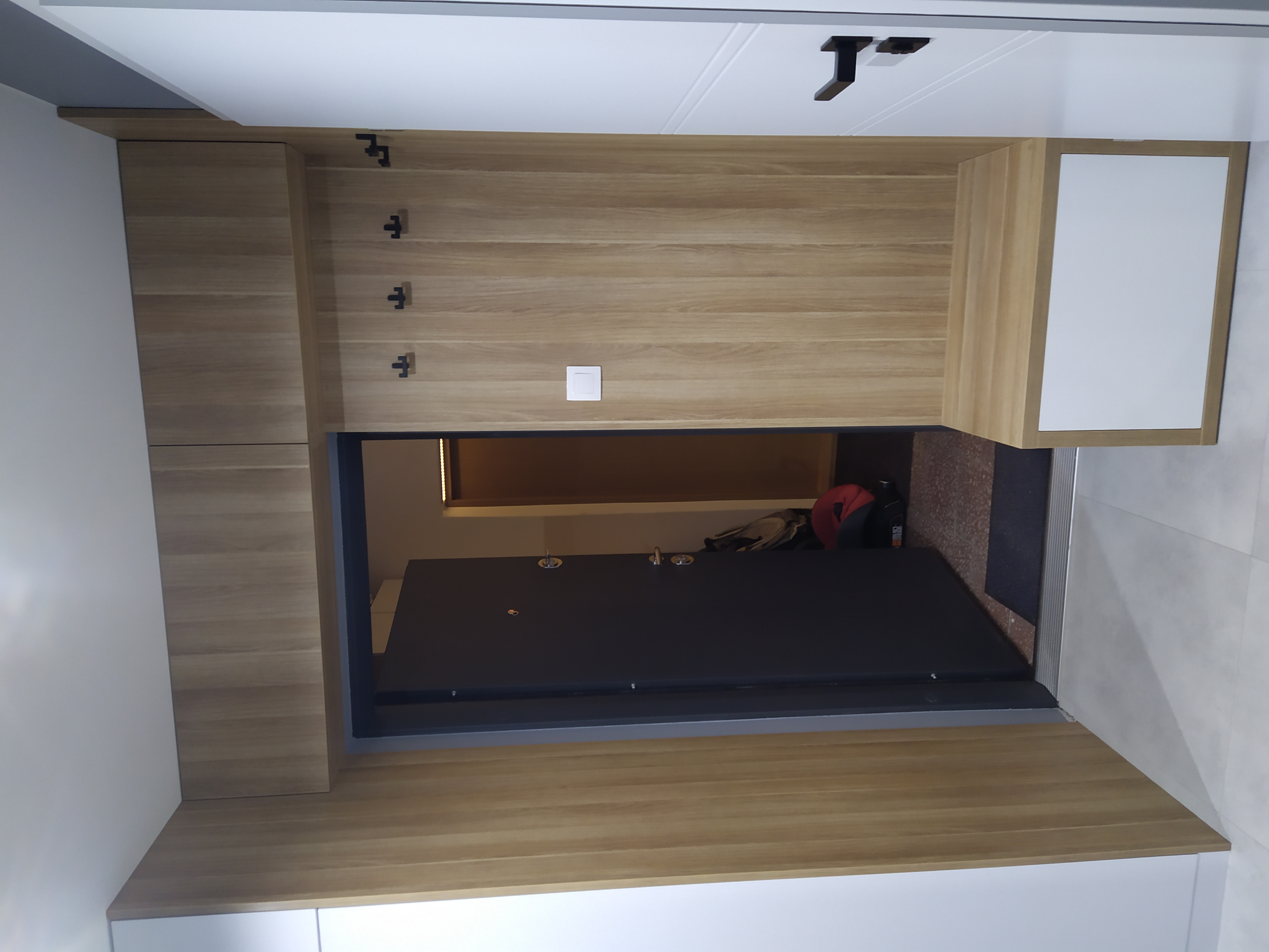 Wnętrze przedpokoju z zabudową z jasnego drewna, w której znajdują się wieszaki na ubrania, szafka z białym frontem oraz widoczne wejście do mieszkania z czarnymi drzwiami.