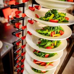 Stojak z białymi talerzami sałatek z rukoli, pomidorów i papryki, przygotowanych do serwowania, z widocznym logo wypożyczalni gastronomicznej w lewym dolnym rogu.