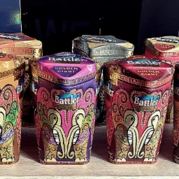 Różnokolorowe, dekoracyjne puszki herbaty Battler Golden Giant, ustawione na drewnianej półce, z logo WG widocznym w tle.