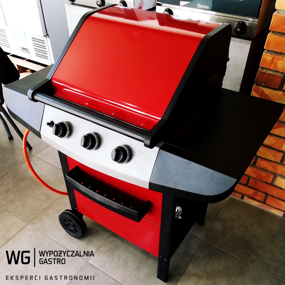 Czerwono-czarny grill gazowy na kółkach z trzema pokrętłami, widoczny w tle fragment ceglanej ściany i sprzętu gastronomicznego.