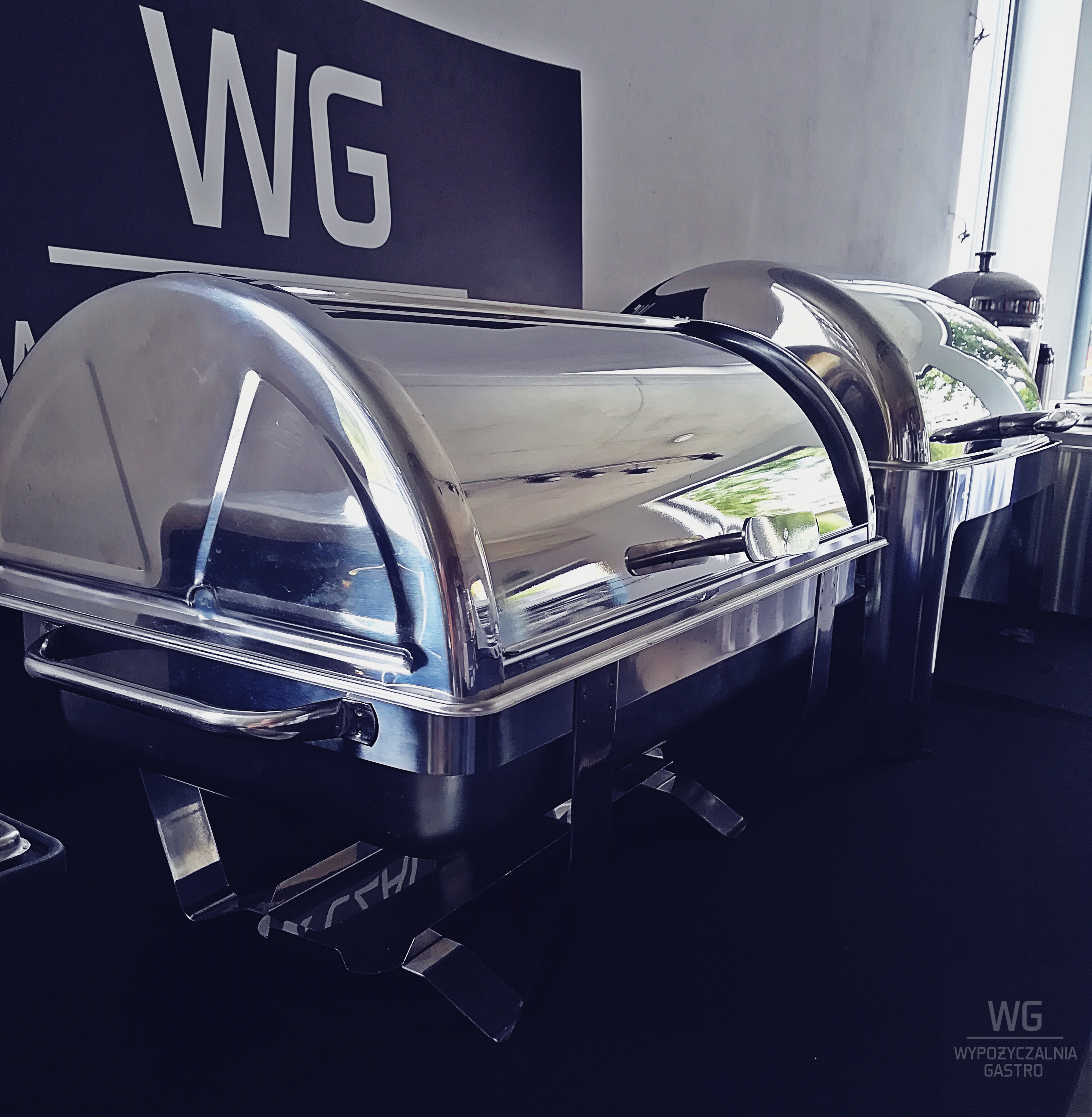Dwa podgrzewacze gastronomiczne ze stali nierdzewnej z przezroczystymi pokrywami, na czarnym stole, z logo firmy WG w tle.