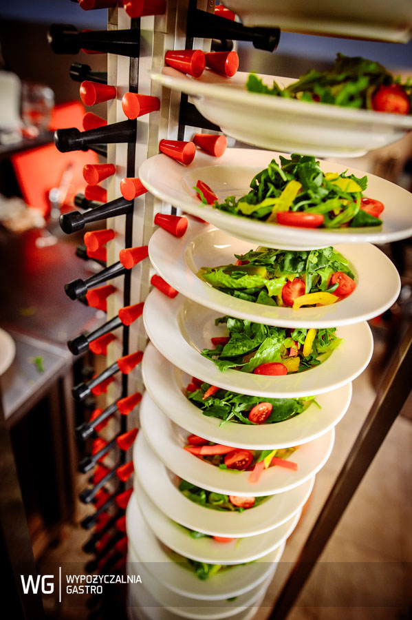 Stojak z białymi talerzami sałatek z rukoli, pomidorów i papryki, przygotowanych do serwowania, z widocznym logo wypożyczalni gastronomicznej w lewym dolnym rogu.