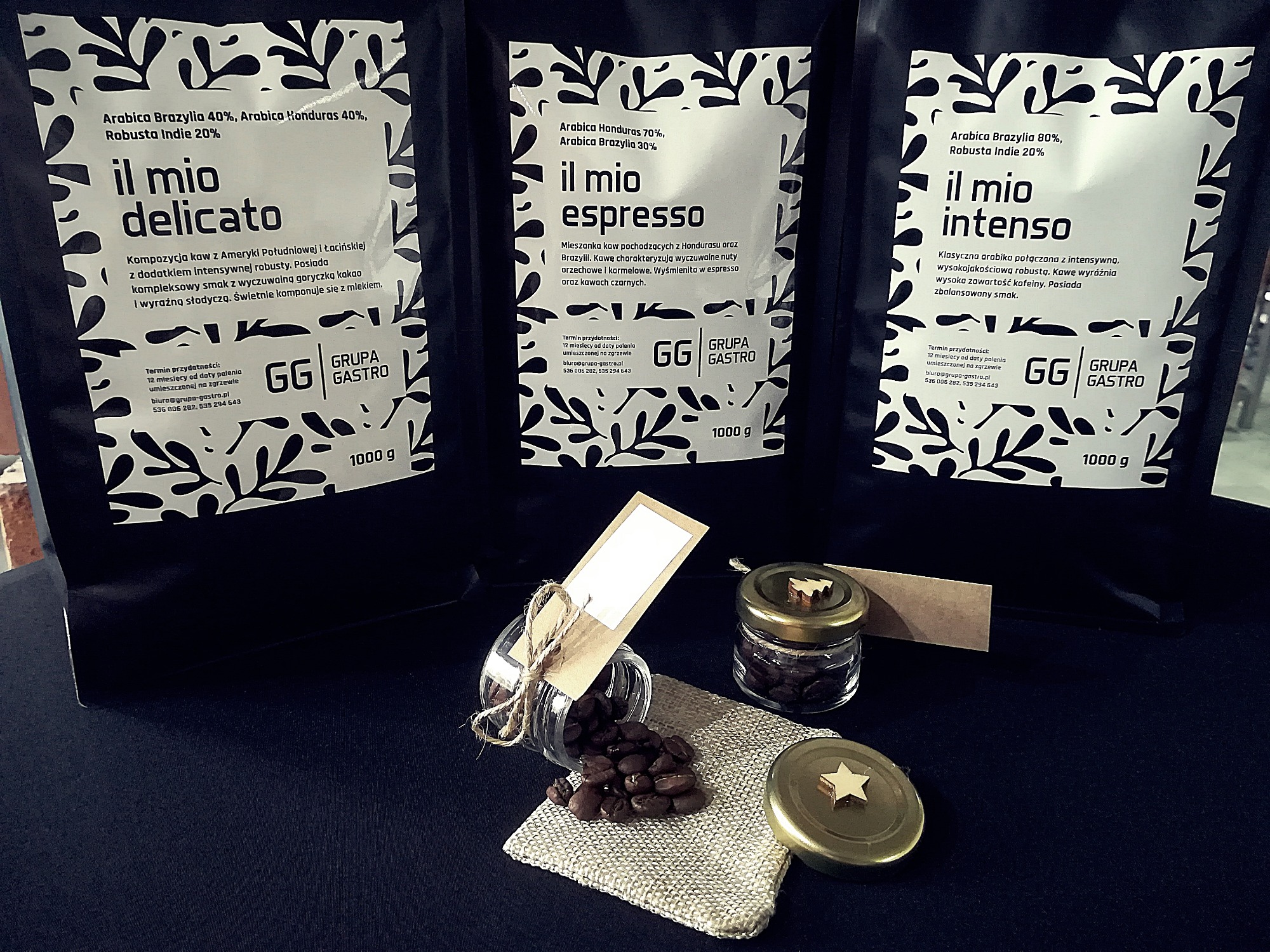 Trzy opakowania kawy ziarnistej 'il mio' o różnych kompozycjach (delicato, espresso, intenso) obok rozsypanych ziaren kawy z małych słoiczków na jutowym podkładzie.