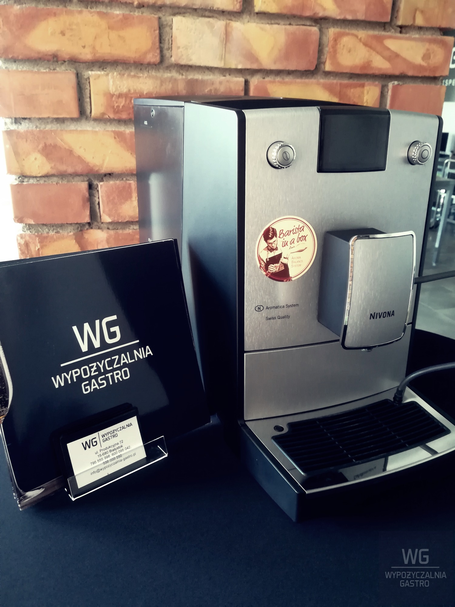 Profesjonalny ekspres do kawy Nivona z naklejką 'Barista in a box' oraz wizytówką firmy WG Wypożyczalnia Gastro na tle ceglanej ściany.