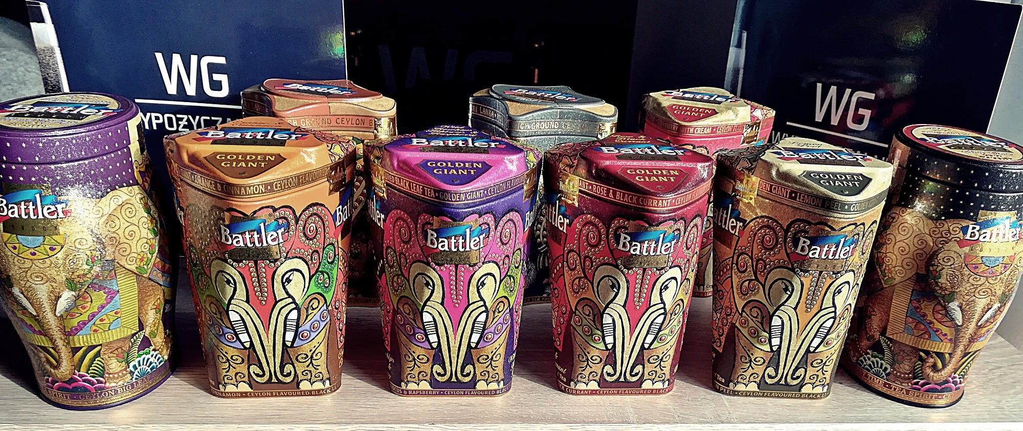 Różnokolorowe, dekoracyjne puszki herbaty Battler Golden Giant, ustawione na drewnianej półce, z logo WG widocznym w tle.