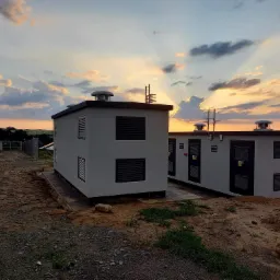 Dwa białe budynki z kratkami wentylacyjnymi i antenami na dachu na tle zachodzącego słońca i paneli słonecznych w oddali.