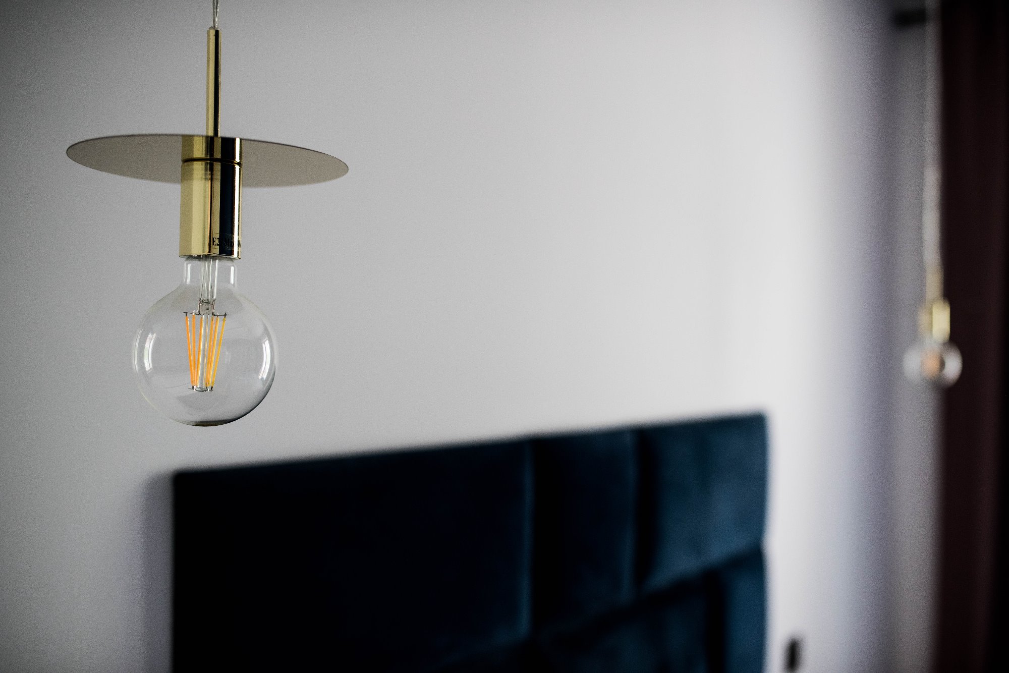Lampa wisząca z żarówką Edison na złotym uchwycie, w tle wezgłowie łóżka obite granatowym welurem.